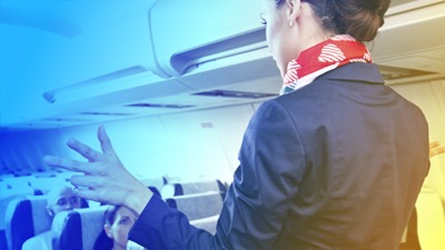 A female flight attendant inside an aircraft talking to the passengers, counting three fingers with her left hand.